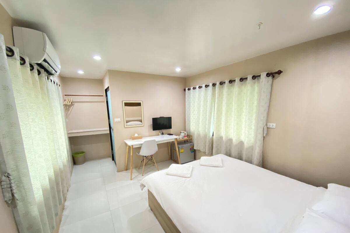 A hotel room with a bed, a desk with a computer, a mirror, and an air conditioning unit.