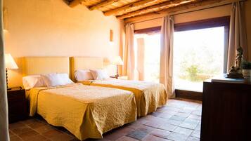 A room with two beds, wooden beams on the ceiling, a large window with curtains, and a nightstand with a lamp.