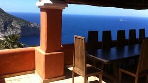 A terrace with a table and chairs, overlooking a coastal view with the sea and a distant boat.