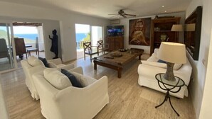 A spacious living room with a white sofa, a coffee table, and a TV. There is a statue, a ceiling fan, and a view of the ocean through large windows.