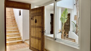 A wooden door with a metal handle, a staircase leading to a white-walled room, and a large window with a view of a plant and a door.