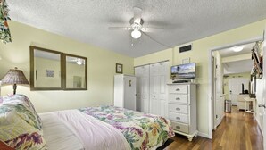 A bedroom with a bed, a dresser, a television, and a ceiling fan. - Sunset Cove: One-Bedroom in Treasure Island (Treasure Island)