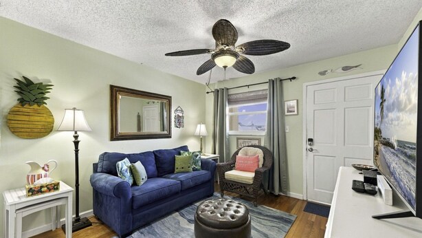 A living room with a blue sofa, a ceiling fan, a television, and a window with curtains. - Sunset Cove: One-Bedroom in Treasure Island (Treasure Island)