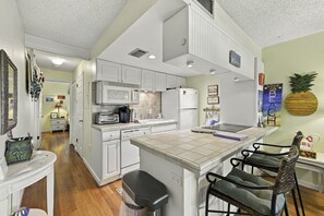 Una cocina con gabinetes blancos, microondas, refrigerador y una pequeña isla con taburetes de bar.