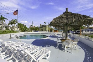 Une piscine avec des chaises longues et un parasol en chaume, entourée de palmiers et surplombée d’un ciel dégagé.