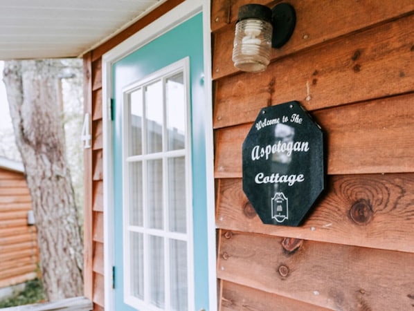Exterior de una cabaña de madera con una puerta color turquesa y un letrero que dice 'Welcome to The Aspotogan Cottage'.