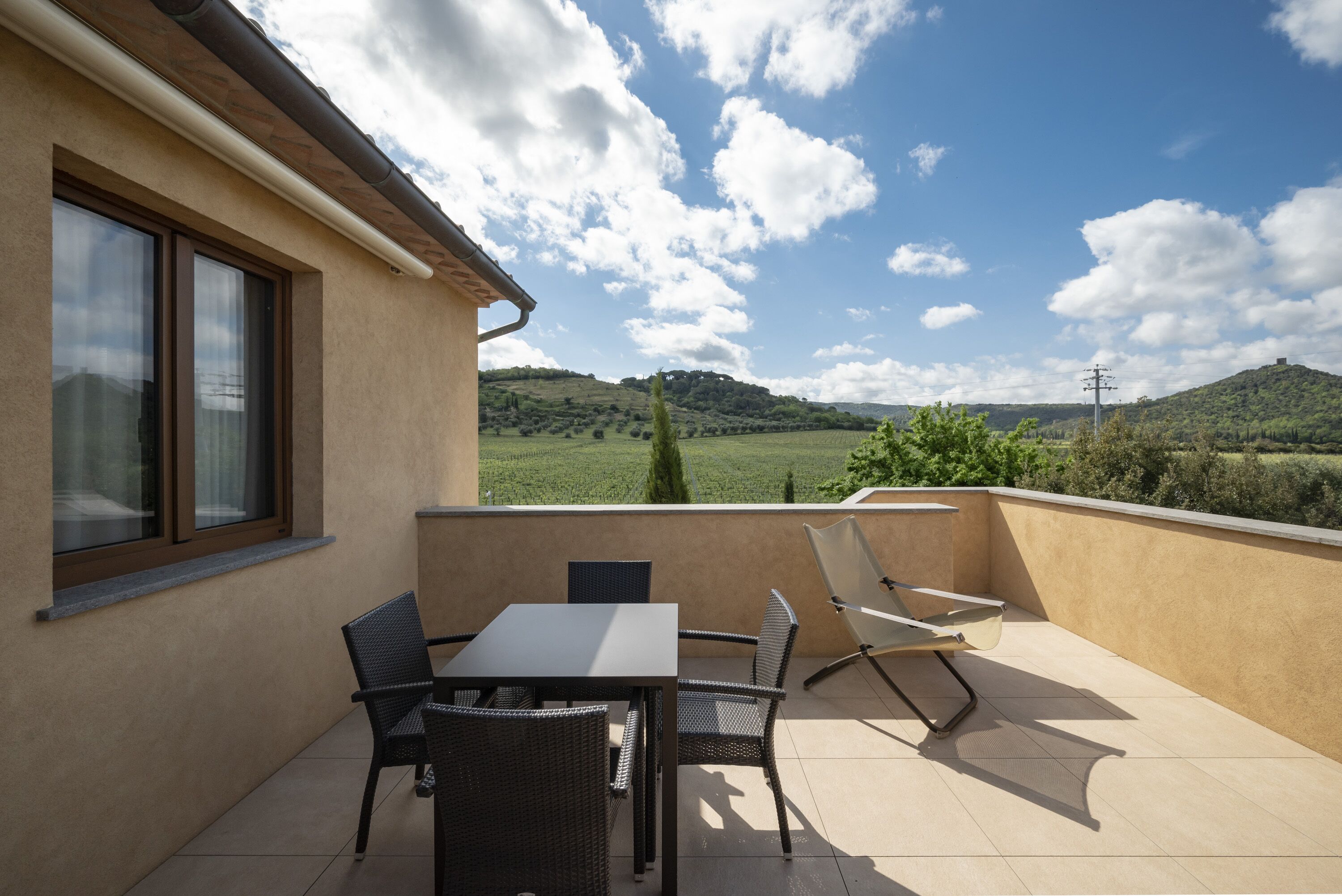 Una terrazza con tavolo e sedie, una finestra e vista su dolci colline verdi sotto un cielo parzialmente nuvoloso.