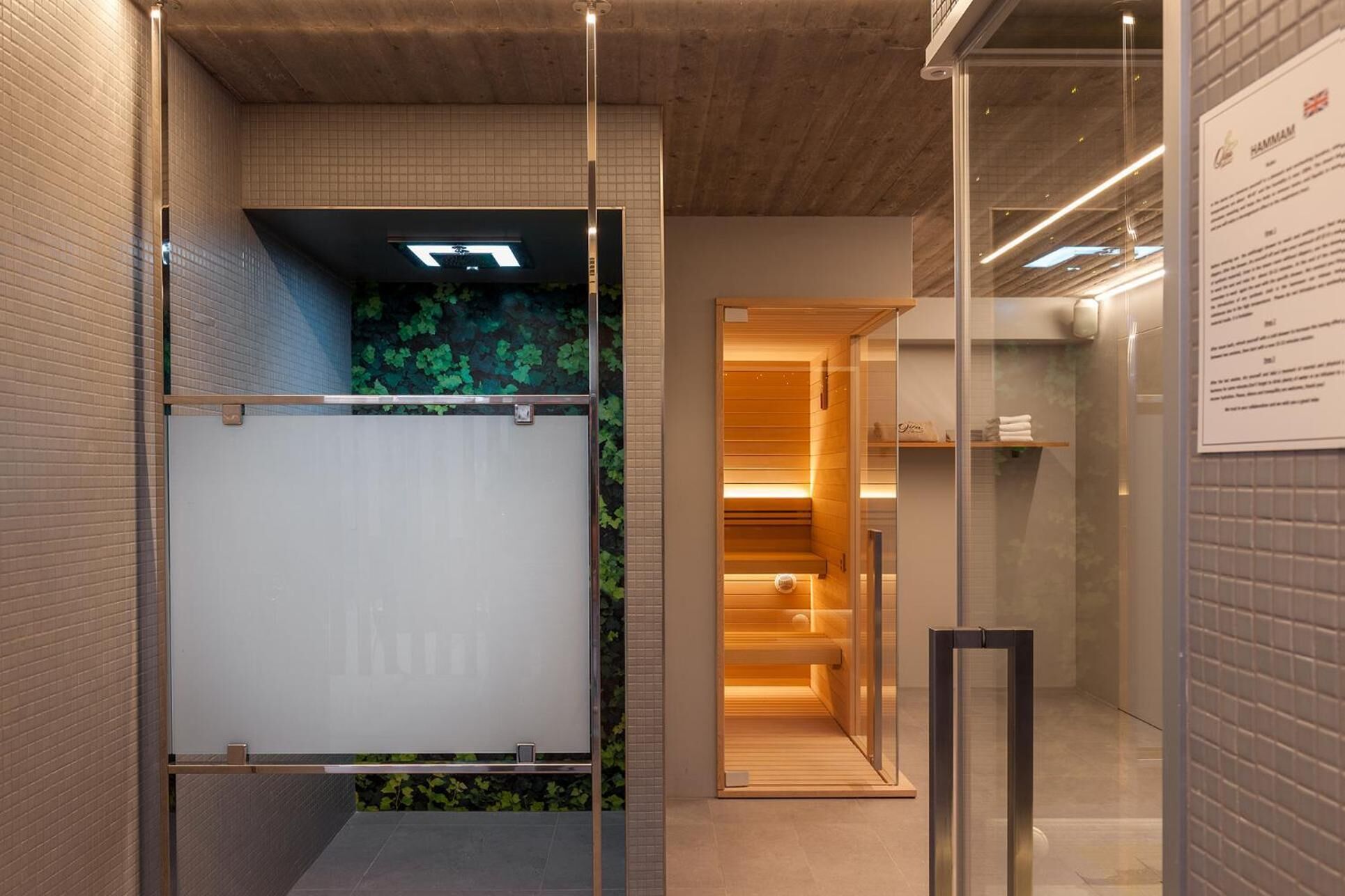 Un interno moderno con una porta in vetro che dà sull'area sauna, una parete rivestita di piastrelle con motivo a griglia e una panca in legno.
