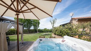 A hot tub on a wooden deck with an umbrella, surrounded by a grassy yard and a building with a tiled roof. - Il Cru di Vignanova (Castagneto Carducci)