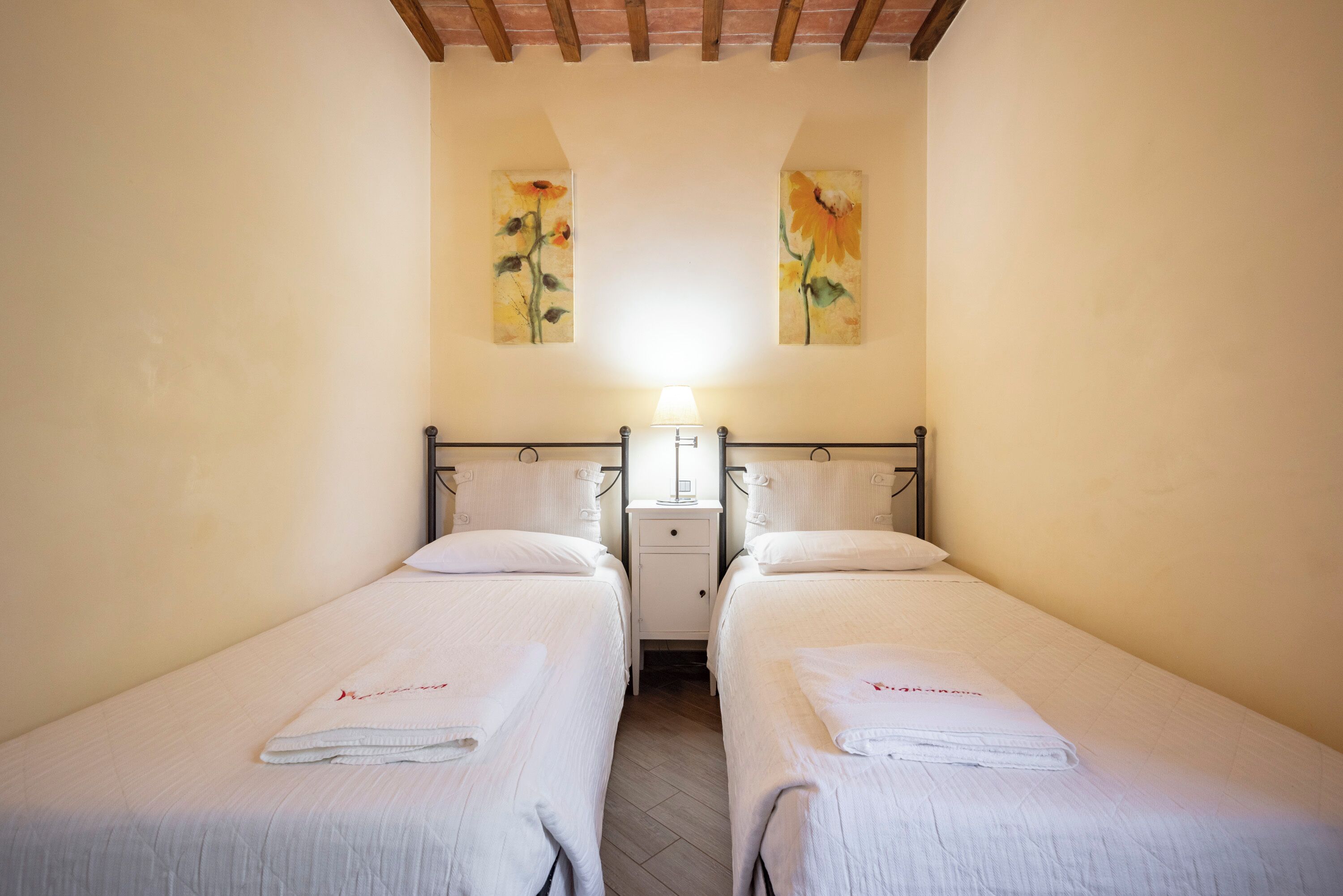 A room with two single beds, a nightstand with a lamp, and two paintings on the wall.