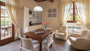 A dining area with a wooden table, white chairs, a vase with flowers, and a kitchen in the background. - Il Cru di Vignanova (Castagneto Carducci)