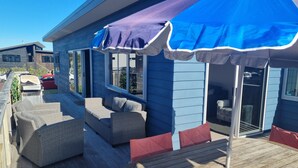 Een terras met rieten meubels en een houten tafel, blauw bekleedde gevel en een overdekt gebied met parasols.