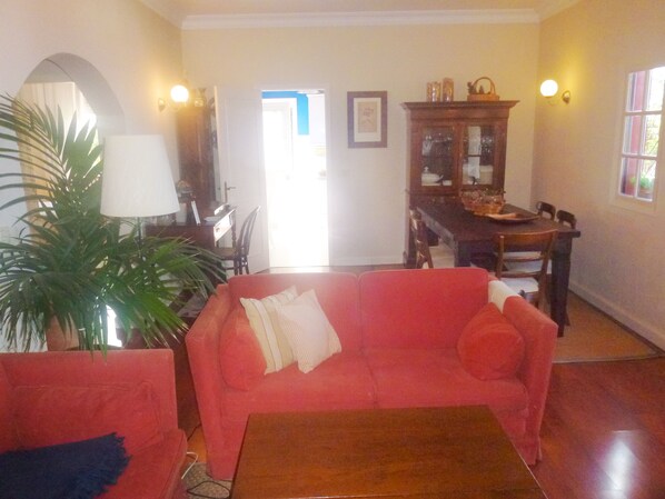 Un salón con un sofá rojo, una mesa de centro de madera y una mesa de comedor con sillas. Hay una lámpara, una planta en maceta y un cuadro en la pared.
