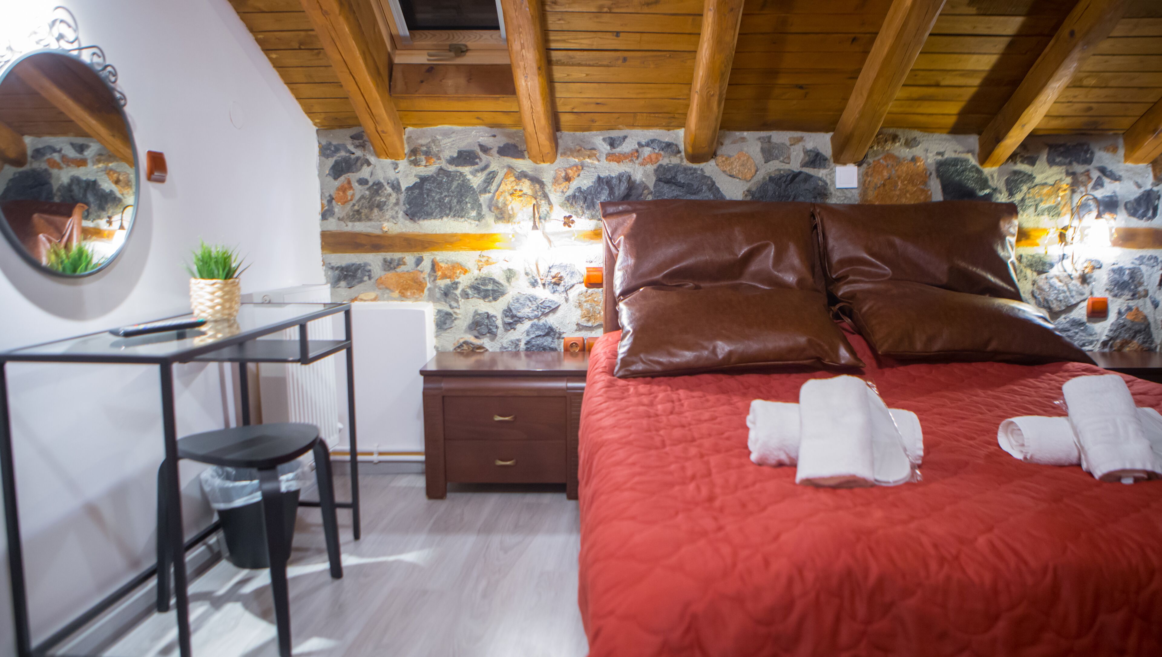 A bedroom with a stone wall, wooden ceiling beams, a bed with red bedding, two pillows, a nightstand, a mirror, a small table with a plant, and a chair.