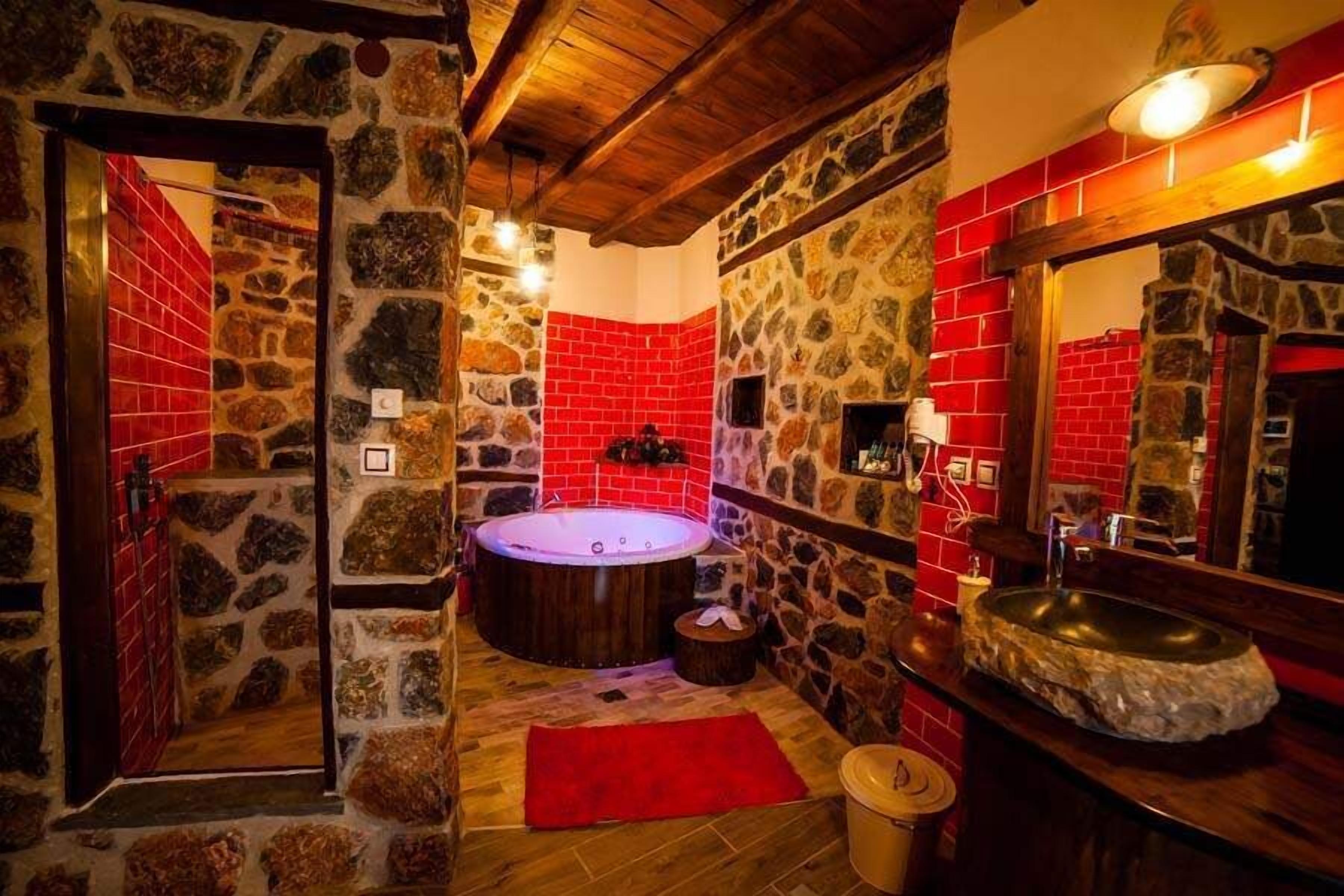A stone-walled bathroom with a red-tiled shower area, a wooden bathtub, and a stone sink.