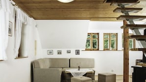 Ein gemütliches Wohnzimmer mit einer Eckcouch, einem kleinen Tisch und einem Kamin. An den Wänden hängen gerahmte Bilder und ein Fenster bietet Blick auf Grünflächen.