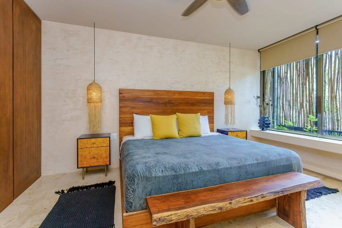 Une chambre à coucher avec une tête de lit en bois, un lit recouvert d’une couverture bleue, deux tables de chevet, un banc et une grande fenêtre donnant sur un jardin de bambous.