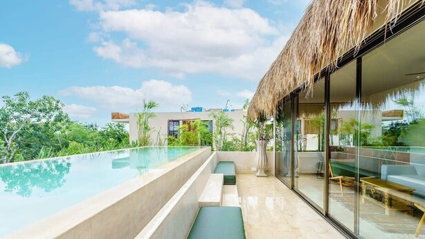 A modern outdoor pool area with a glass enclosure, seating, and a thatched roof structure. - Aldea Zama Condo w/ Private Rooftop Infinity Pool (Tulum)
