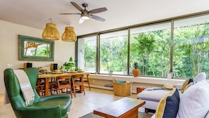 A modern living room with a green chair, a wooden coffee table, and a sofa with cushions. There is a dining table with chairs, a large window with a view of greenery, and a ceiling fan. - Aldea Zama Condo w/ Private Rooftop Infinity Pool (Tulum)