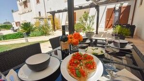 Un espace de restauration en plein air avec une table dressée pour deux personnes, où l’on peut déguster un repas composé de pain, de tomates et de basilic, accompagné de verres à vin et d’une bouteille de vin.