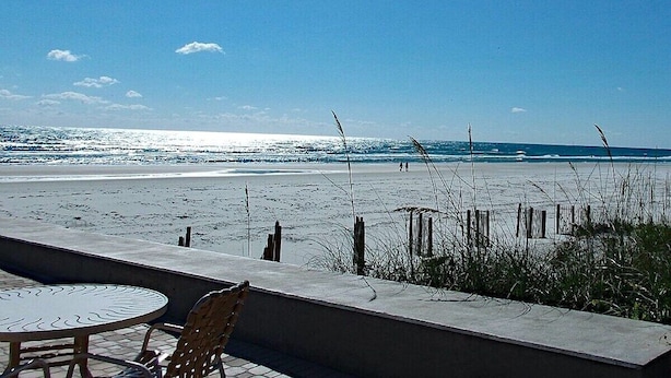 A beach view with a table and chairs, a concrete wall, and a wooden fence. - 402 - Fourth floor ocean view delight (New Smyrna Beach)