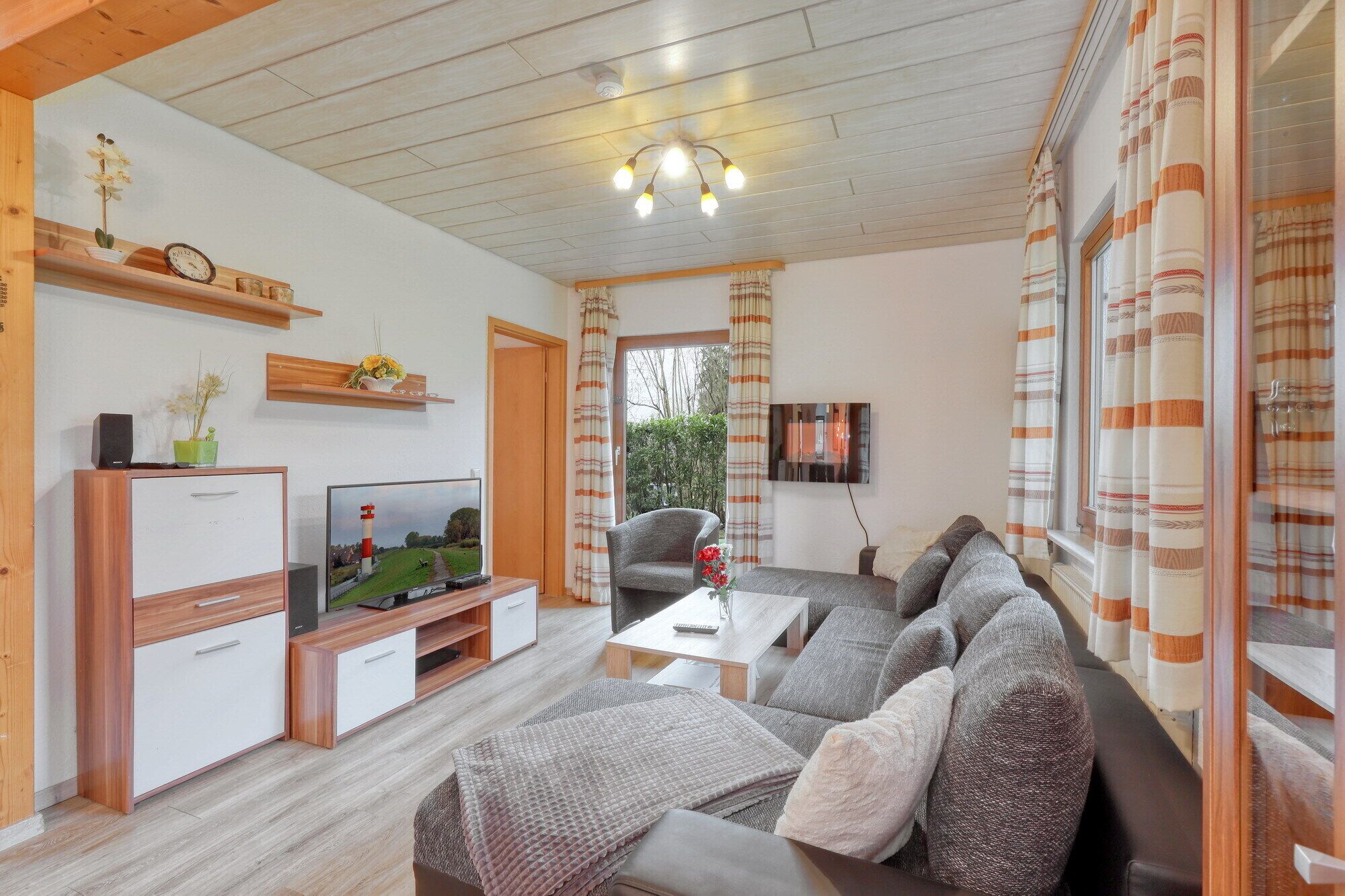 A living room with a sofa, TV, and a small table. There is a wooden cabinet with a plant and a clock on top. The room has a wooden ceiling and curtains on the windows.