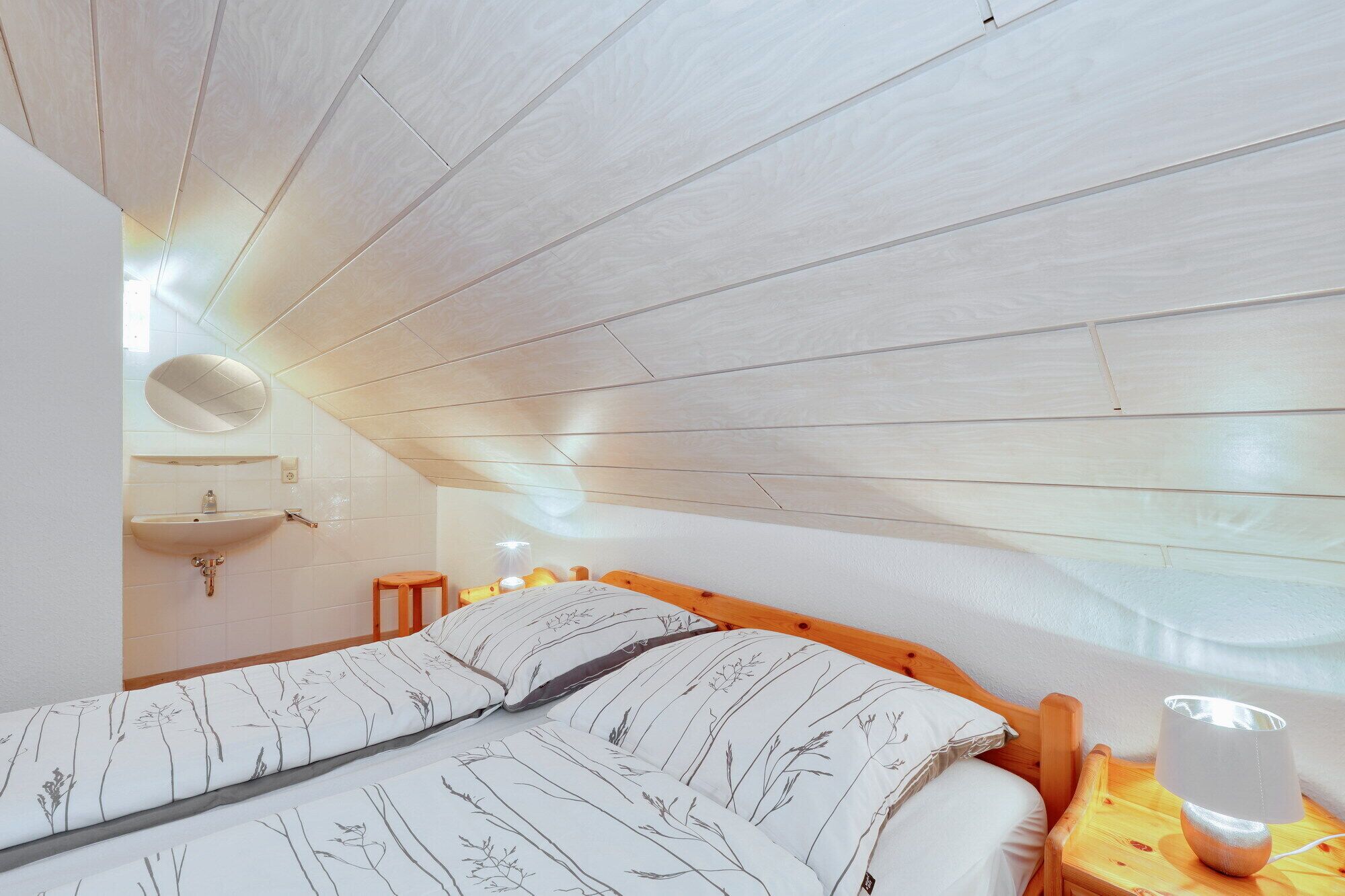 Un dormitorio con techo inclinado, estructura de cama de madera, mesita de noche con lámpara y espejo montado en la pared.