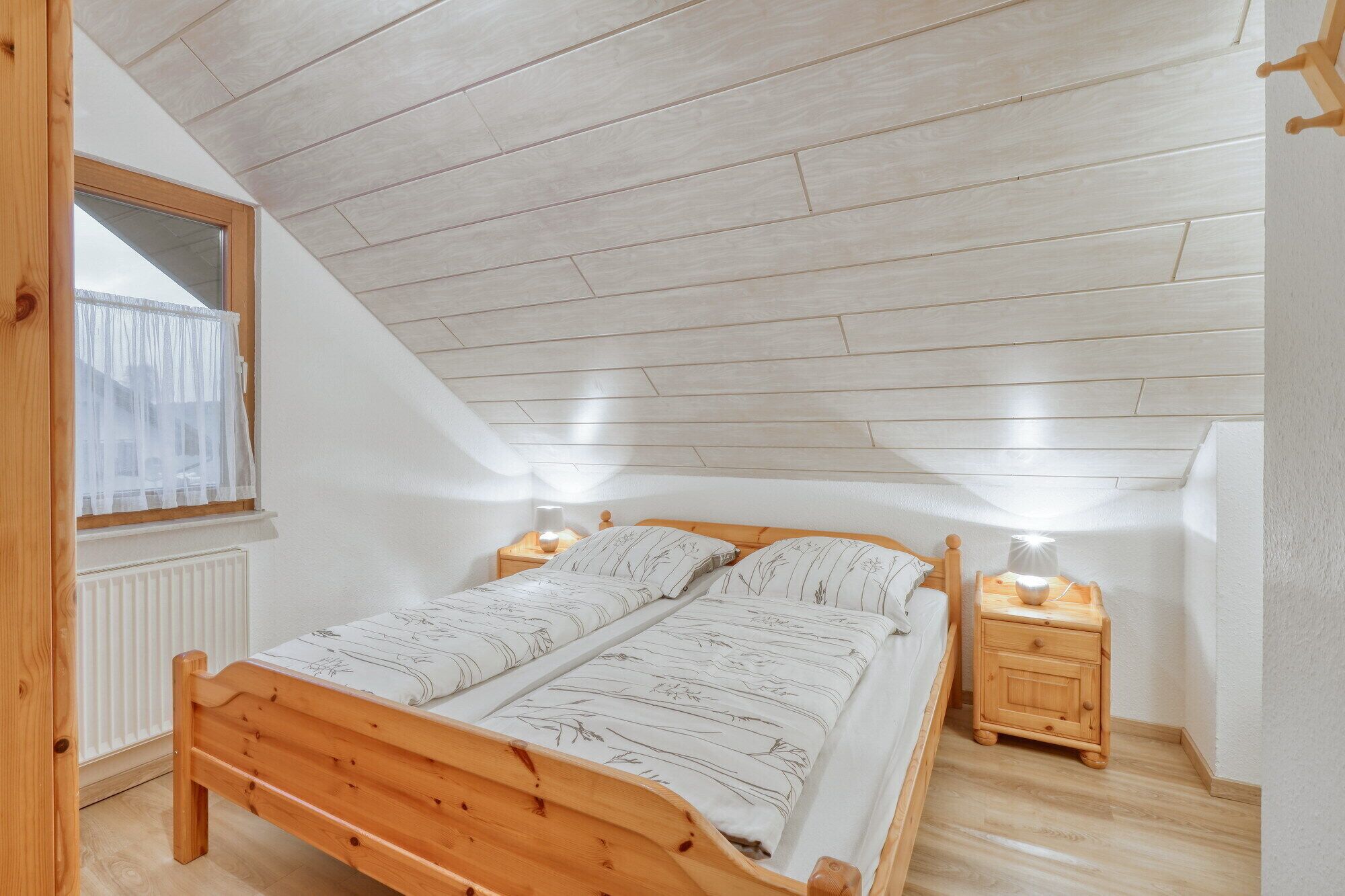 A bedroom with a sloped ceiling, wooden bed frame, two bedside tables with lamps, and a window with a sheer curtain.