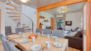 Uma sala de jantar com uma mesa de madeira posta para quatro pessoas, rodeada por cadeiras. Uma escadaria é visível à esquerda e uma sala de estar com sofá e televisão ao fundo.