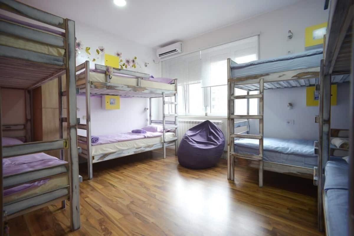 A dormitory room with bunk beds, wooden flooring, an air conditioning unit, and a window.