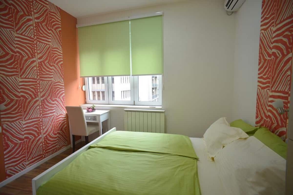 A bedroom with a bed, a desk, and a window with blinds.