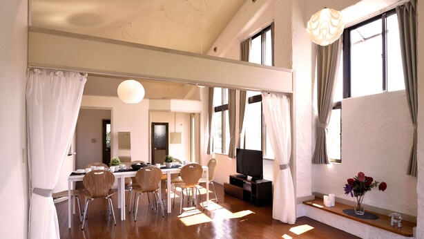 A spacious room with a wooden floor, a dining area with chairs and a table, a TV on a stand, and a shelf with a vase of flowers. - Bay Side Okinawa (Onna)