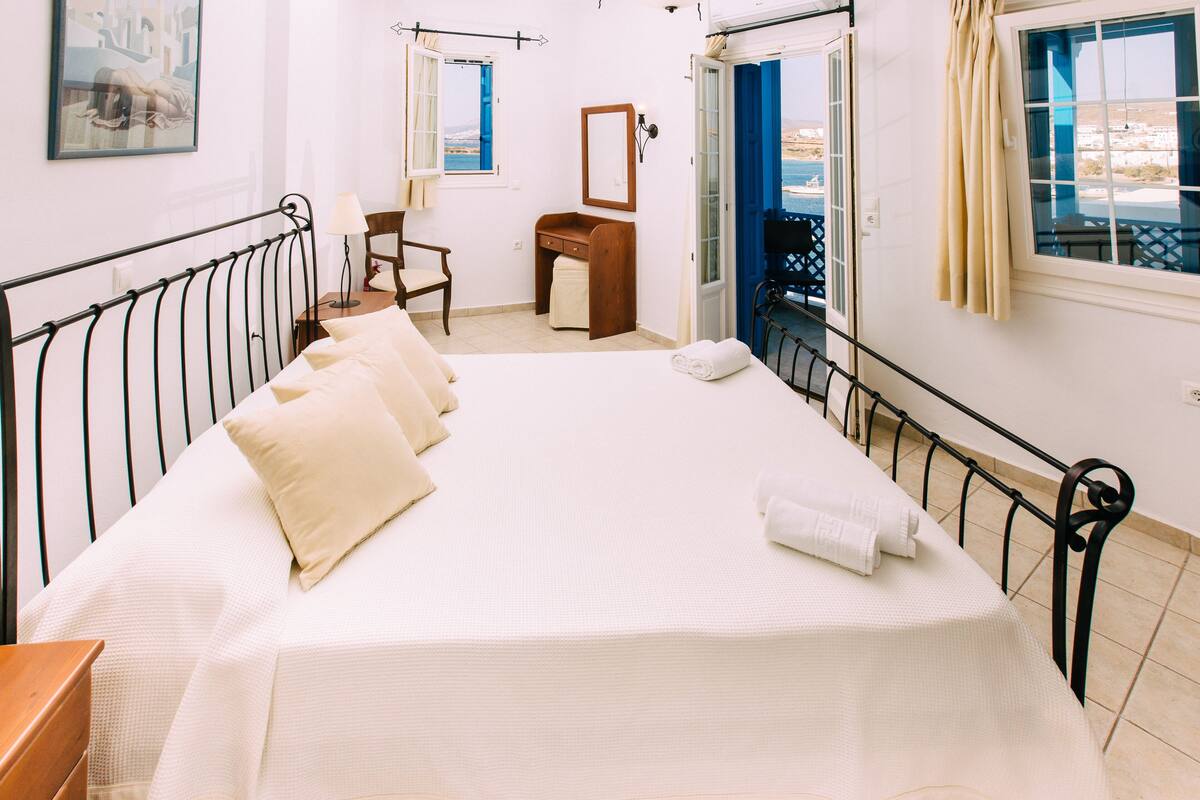 A bedroom with a bed, a desk, a chair, a mirror, and a view of the sea through the window.