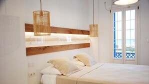 Et soveværelse med sengegavl i træ, seng med hvidt sengetøj, to sengeborde med lamper, og havudsigt gennem en dør.