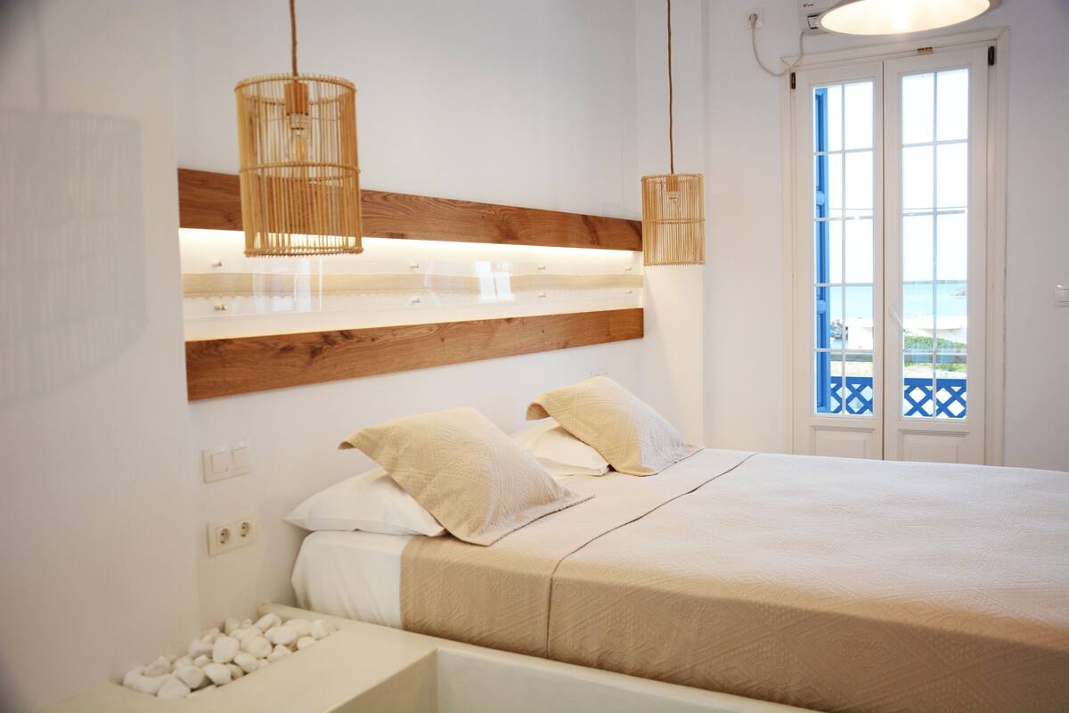 A bedroom with a wooden headboard, a bed with white bedding, two bedside lamps, and a view of the sea through a door.