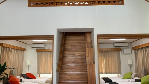 A hotel room with a wooden staircase leading to a balcony, two beds with white bedding and colorful pillows, and a wooden ceiling design. - Nomu House (Chatan)