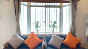 A room with a large window, two wicker sofas with cushions, and two potted plants. - Nomu House (Chatan)