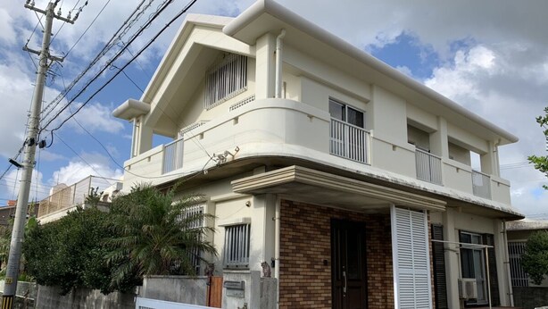 A two-story residential building with a balcony, a brick entrance, and a gated driveway. - Nomu House (Chatan)