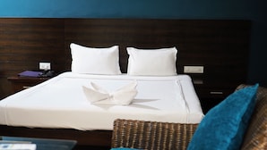 Una cama bien hecha con sábanas y almohadas blancas, y un cojín azul sobre la cama, junto con una silla de mimbre con un cojín azul.