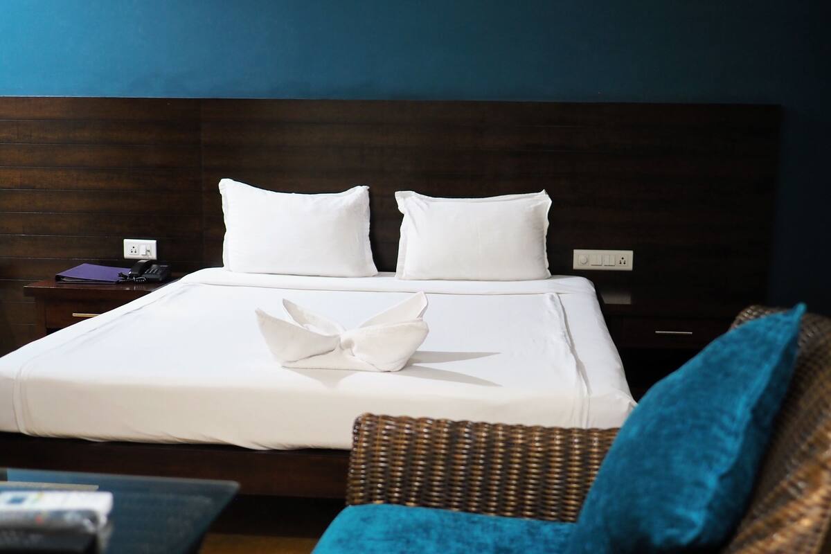 Een net opgemaakt bed met witte lakens en kussens, een blauw kussen op het bed, en een rieten stoel met een blauw kussen.