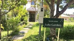 Skilt der byder besøgende velkommen til Lulu Garden, en sti der fører hen til en bygning med et gult tag, omgivet af frodig vegetation.