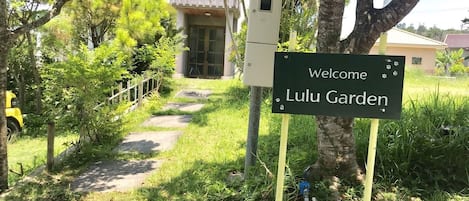 Un letrero da la bienvenida a visitantes a Lulu Garden, un camino que lleva a un edificio con techo amarillo, rodeado de exuberante vegetación.