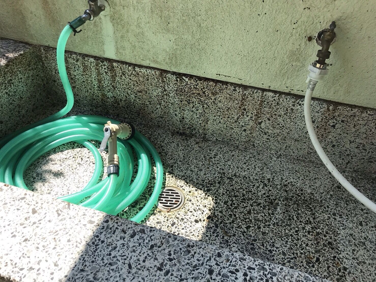 Una manguera verde enrollada sobre un suelo de piedra moteada, cerca de una pared con una rejilla de desagüe.