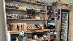 A rustic café interior with wooden tables and chairs, shelves stocked with coffee beans and pastries, and a refrigerator with beverages.