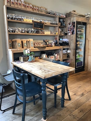 A rustic café interior with wooden tables and chairs, shelves stocked with coffee beans and pastries, and a refrigerator with beverages.