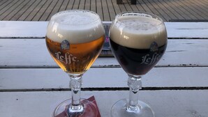 Two beer glasses on a white wooden table with a view of a deck area where people are seated at tables.