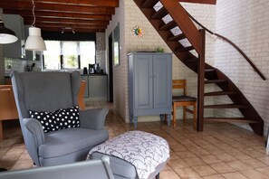 Un salon chaleureux avec un canapé gris, un pouf à motifs, une chaise en bois, un meuble gris et un escalier en bois adossé à un mur de briques blanches.