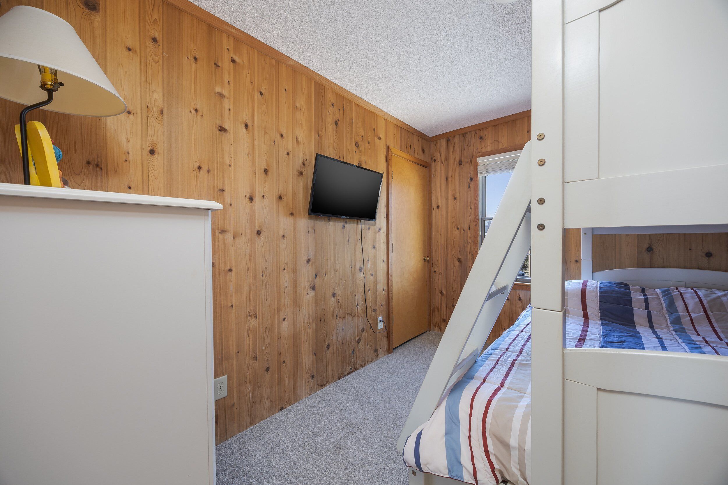 A bedroom with a bunk bed, a television mounted on the wall, and a lamp on a dresser.