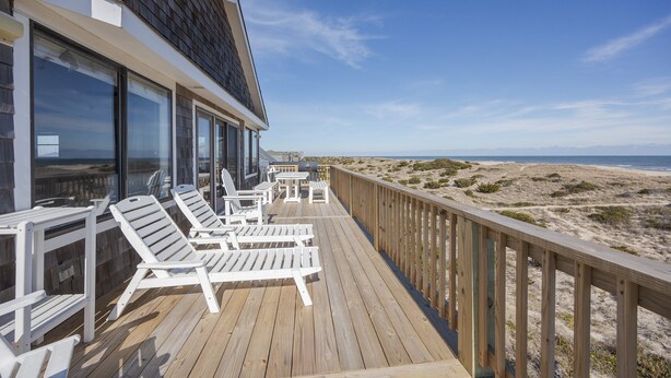 Une terrasse en bois avec des chaises longues blanches et une table, donnant sur une plage bordée de dunes.