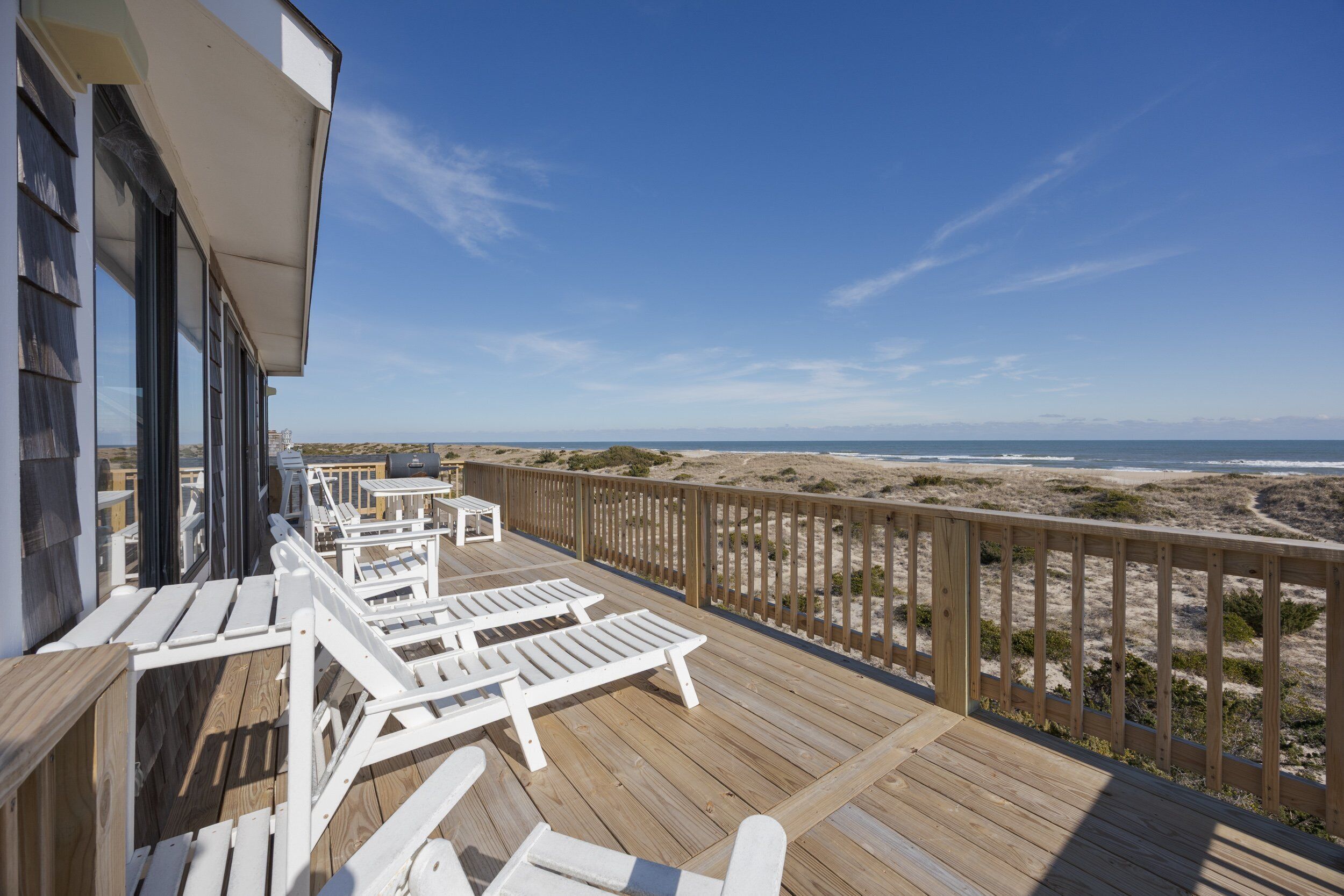 A wooden deck with white lounge chairs and a table, overlooking a beach with dunes and the ocean.